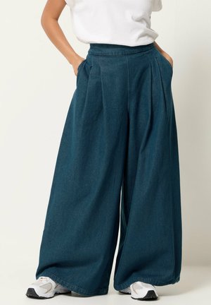 Person iført brede mørk denim bukser med forreste læg, hvide sneakers og en hvid t-shirt stukket ned, hænderne i lommerne.