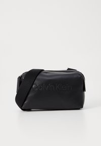 Černá crossbody taška z umělé kůže se zipovým uzávěrem, zdobená embosovaným logem "Calvin Klein", s nastavitelným popruhem a hladkým povrchem.