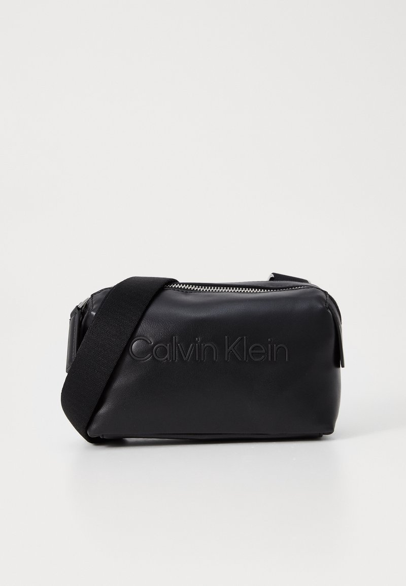 Černá crossbody taška z umělé kůže se zipovým uzávěrem, zdobená embosovaným logem "Calvin Klein", s nastavitelným popruhem a hladkým povrchem.