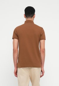 Vīrietis ar īsiem lokainiem matiem, ģērbies brūnā īsām piedurknēm polo krekliņā un bēšās biksēs, redzams no aizmugures uz vienmuļa fona.