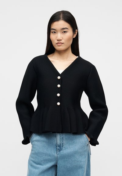 Γυναίκα με μακριά σκούρα μαλλιά, που φοράει μαύρη πλεκτή ζακέτα με πέρλες κουμπιά και ανοιχτόχρωμο τζιν, στέκεται μπροστά σε έναν απλό φόντο.