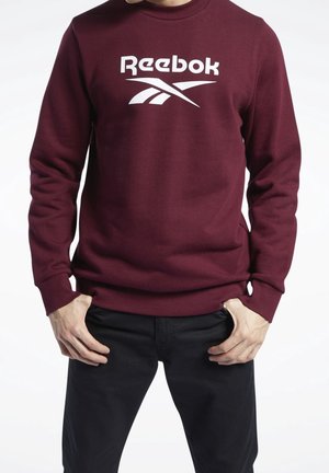 Person trägt einen burgunderfarbenen Reebok-Sweatshirt mit weißem Logo, kombiniert mit schwarzen Hosen, steht mit den Händen in den Taschen auf weißem Hintergrund.
