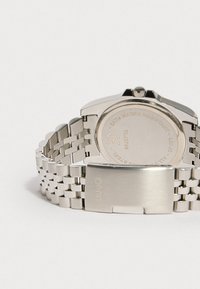 Ασημένιο ρολόι από ανοξείδωτο ατσάλι με βραχιόλι συνδεδεμένων κομματιών, στρογγυλή κάσα και αδιάβροχη πλάτη που δείχνει σκαλιστές λεπτομέρειες και λογότυπο.