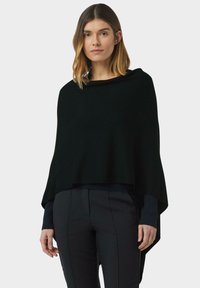 Donna con capelli di lunghezza fino alle spalle, che indossa un poncho nero su una maglietta a maniche lunghe e pantaloni neri, che si trova davanti a uno sfondo bianco uniforme.