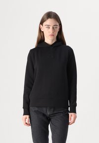 Sweat à capuche noir avec une texture douce et lisse, un logo subtil sur le devant et des poignets et un ourlet côtelés. Associé à un jean sombre.
