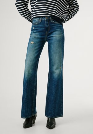Jeans a zampa in denim blu con dettagli strappati. Presentano una vita aderente e una leggera sfumatura, abbinati a stivali neri con tacco texture.