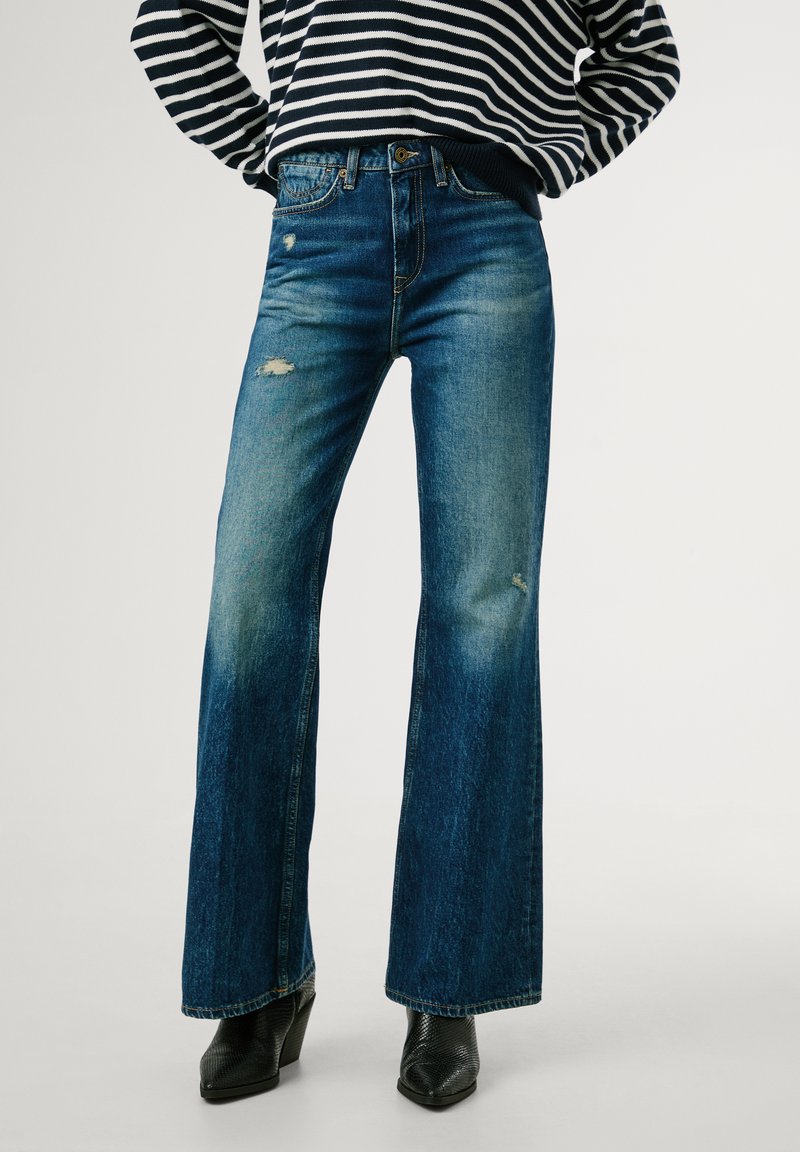 Uitlopende blauwe spijkerbroek van denim met versleten details. Heeft een aangesloten taille en een lichte vervaging, gecombineerd met gestructureerde zwarte haklaarzen.
