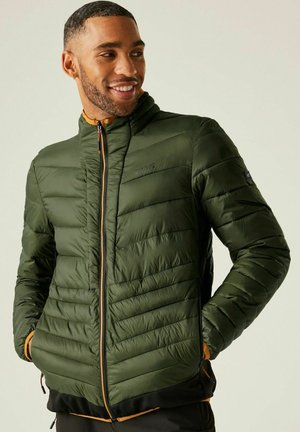 Uśmiechnięty mężczyzna w zielonej pikowanej kurtce z zamkami błyskawicznymi w kieszeniach, z rękami w kieszeniach, patrzący na bok na jasnym, jednolitym tle.