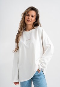 Felpa bianca a maniche lunghe con scollo tondo e taglio morbido, con la scritta "MIRONS" stampata in un sottile rosa sul petto. Abbinata a jeans azzurri chiari.