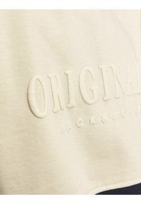 Tela crema con texto elevado "ORIGINALS" y "JACK & JONES" más pequeño debajo en el material de la ropa.