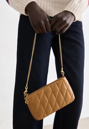 Mains tenant un sac à main en cuir beige matelassé avec une bandoulière chaîne dorée, posé sur un pantalon foncé et un pull crème.