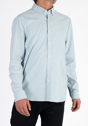 Mand iført lyseblå skjorte med knapper og lange ærmer samt krave og sorte bukser, stående mod hvid baggrund.