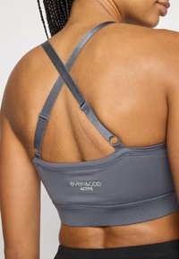 Šedá sportovní podprsenka s překříženými ramínky, vyrobená z elastické látky. Na lemu je logo a nastavitelné přezky na ramínkách. Hladký povrch.