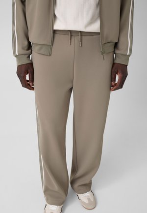 Persona con pantalones deportivos color topo con cordón, chaqueta a juego con rayas blancas en las mangas, camiseta blanca y zapatillas blancas.