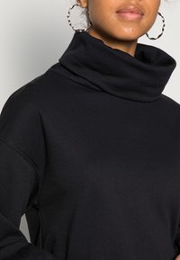Sweat-shirt noir à col roulé en tissu doux, coupe décontractée et col plié. Des boucles d'oreilles anneaux argentées accentuent le look.