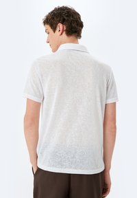 Hvid polo skjorte med en tekstureret overflade, korte ærmer og en klassisk krave. Har et let gennemsigtigt design på ryggen.
