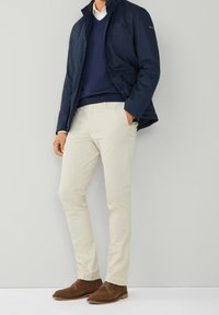 Marinblå jacka, blå tröja och beige byxor. Bruna mockaskor. Jackan har en ståkrage och bröstficka. Släta texturer genomgående.