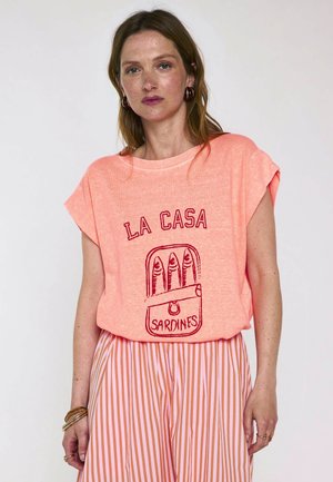 Femme portant une chemise couleur pêche avec "La Casa Sardines" et un motif de boîte de sardines, associée à une jupe à rayures orange et blanches, debout sur fond blanc.
