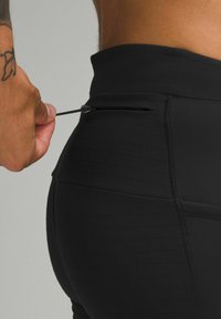 Fekete sportleggings, rácsos mintával, magas derékkal és diszkrét cipzáros zsebbel. A textúra sima, kiegészítő varrási részletekkel.