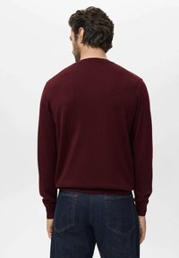 Man wearing a burgundy long-sleeve sweater and dark blue jeans facing away against a plain light background.
