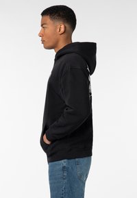 Schwarzer Baumwollhoodie mit Kängurutasche, gerippten Bündchen und Kapuze, auf dem Rücken mit einem grafischen Druck. Getragen mit blauen Jeans.