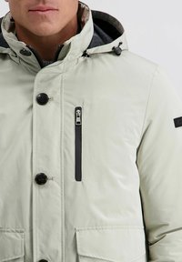 Man in een lichtgrijze winterjas met capuchon, zwarte knopen, een verticale ritszak op de borst en klepzakken aan de voorkant.