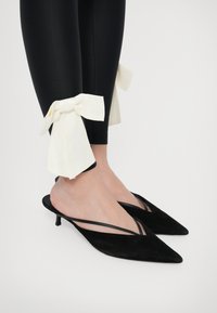 Mustast siidist slingback-kontsad, millel on terav nina ja õhuke rihma disain. Kombineeritud mustade retuusidega, millel on valged kangast paelad varrukatel.