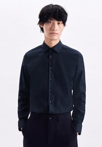 Camicia lunga blu navy con vestibilità slim, colletto button-down e bottoni coordinati. Il tessuto appare liscio con una leggera lucentezza. Pantaloni slim.