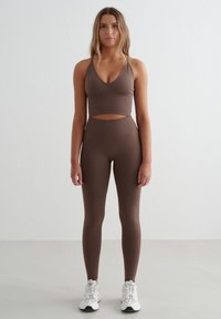 Bruine sportbeha en high-waisted leggings in rekbare, textuurstof. De beha heeft een uitsnijding bij de taille. Witte sneakers.