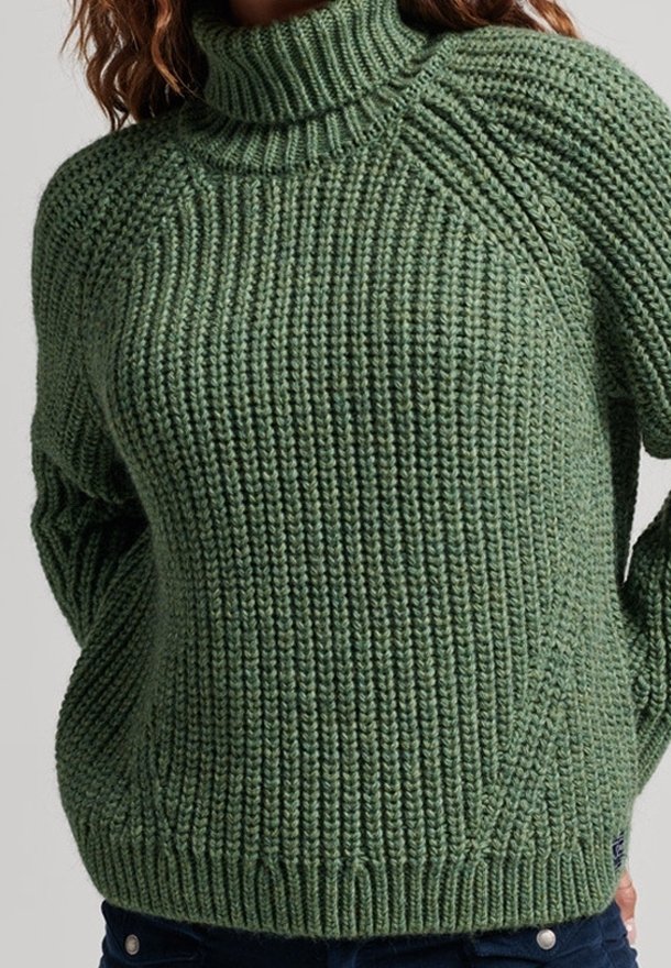 Pull à col roulé vert en grosse maille porté par une personne avec les mains derrière le dos, montrant un motif côtelé texturé.