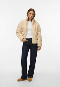 Beige fakepälsjacka med hög krage och två framfickor, bärs över en vit topp och mörkblå jeans, kombinerad med bruna träskor.