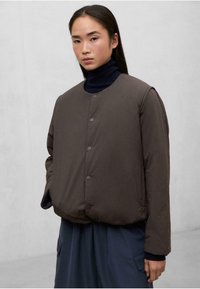 Chaqueta acolchada marrón con cuello redondo, botones a presión y una textura suave. Llevada sobre un suéter oscuro de cuello alto.