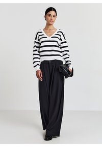 Frau in einem schwarz-weiß gestreiften V-Ausschnitt-Pullover, mit weit geschnittenen schwarzen Hosen, schwarzen Schuhen, die eine schwarze Clutch hält, vor einem einfarbigen Hintergrund stehend.
