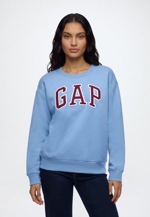 Jeune femme aux longs cheveux foncés portant un sweat-shirt bleu clair avec des lettres "GAP" rouges et blanches et un jean foncé sur un fond uni.