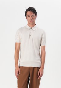 Beige strikket polo-skjorte med korte ermer, tre knappelukking, ribbestrikket krage og hem. Kombinert med brune bukser.