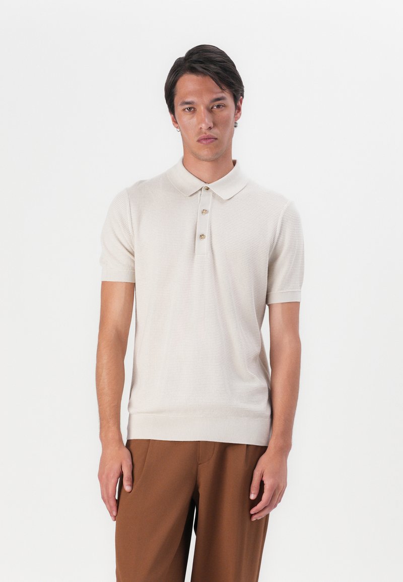 Beige strikket polo-skjorte med korte ermer, tre knappelukking, ribbestrikket krage og hem. Kombinert med brune bukser.