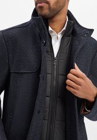 Marineblauer Mantel mit Fischgrätmuster, Stehkragen und Knopfdetails, darüber getragen eine schwarze, gesteppte Innenjacke mit Reißverschluss.