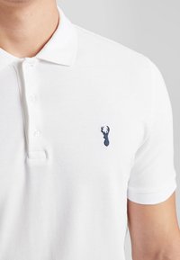 Hvid polo shirt lavet af tekstureret stof med krave, der har et lille marineblåt hjortelogo på venstre bryst. Fire knaplukninger.
