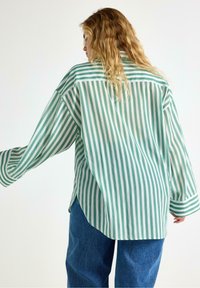 Persona bionda che indossa una camicia oversize a righe verdi e bianche e jeans blu, girata leggermente di lato su uno sfondo neutro.