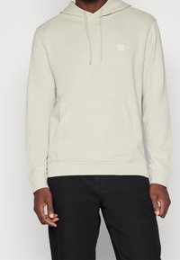 Beige huvtröja med ficka fram, ribbade mudd och nederkant. Har en liten logotyp på bröstet. Materialet verkar mjukt och texturerat.