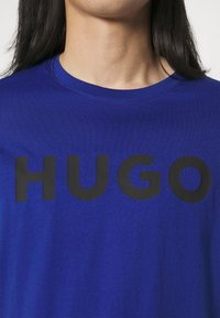 Modrá bavlněná tričko s velkým černým potiskem "HUGO" na hrudi. Kulatý výstřih a hladká textura s volným střihem.