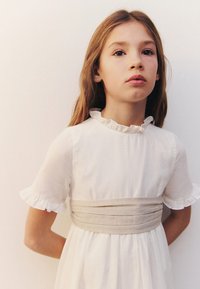Biele šaty s krátkymi volánovými rukávmi, so béžovým opaskom v páse. Tkanina vyzerá ľahko a hladko, s jednoduchým dizajnom.
