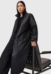 Zwarte waterdichte trenchcoat met een gestructureerde afwerking, voorzien van een hoge kraag en zijzakken, gecombineerd met een zwarte gebreide trui en wijde broek.