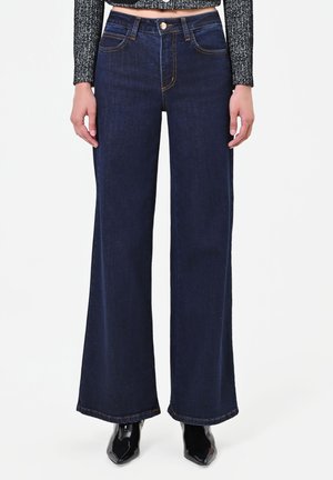 Femme portant un jean large bleu foncé associé à un pull court noir et blanc et des chaussures noires à bout pointu sur fond blanc.
