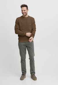 Felpa marrone con collo tondo, polsini a coste e tasca laterale. Abbinata a pantaloni verde chiaro e calzature grigie. Tessuto morbido e liscio.