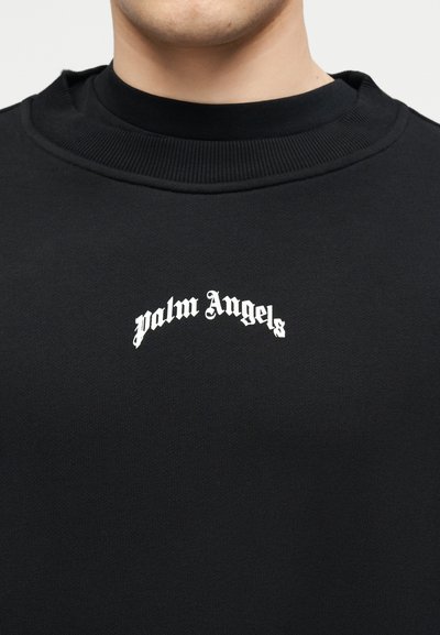 Άνδρας που φοράει μαύρο φούτερ με λευκό κείμενο «palm Angels» σε γοτθική γραμματοσειρά στο κέντρο του στήθους.