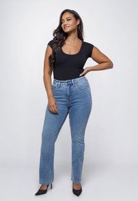 Vrouw met lang, donker haar, gekleed in een strakke zwarte top, lichtblauwe high-waisted jeans en zwarte hakken, staand met de hand op de heup.