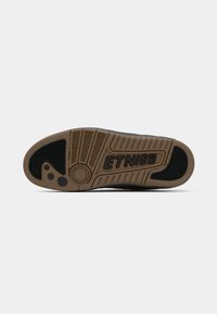 Suela de zapato de patinaje negra con una suela de goma que presenta un patrón de espiga, el logo de Etnies y acentos circulares en tono beige y gris oscuro.