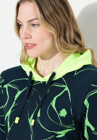 Marine blauwe hoodie met groene abstracte patronen, voorzien van een neon groene voering in de capuchon, verstelbare koordjes en zilveren oogjes.