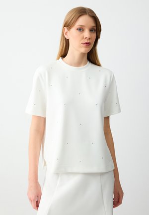 Top bianco a maniche corte con scollatura rotonda, caratterizzato da piccole decorazioni nere su tutto il tessuto e spacchi laterali per facilitare i movimenti.